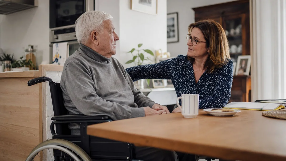 Homme âgé en fauteuil roulant accompagné par sa fille à domicile, illustration du vieillissement avec un handicap